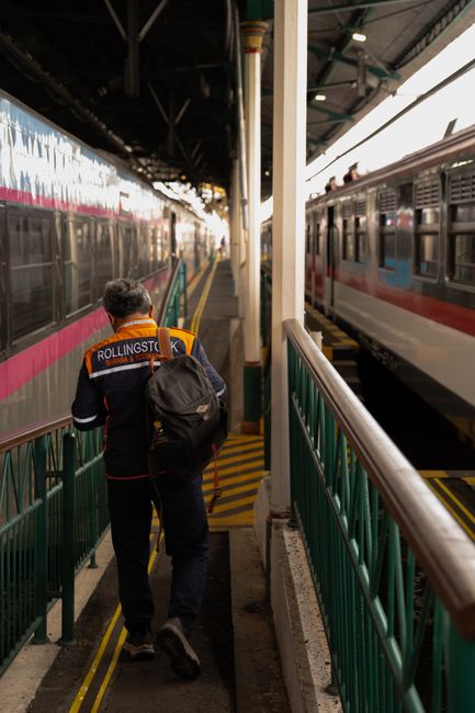 A person wearing a dark blue and orange jacket with 'ROLLINGSTOCK' written on the back, carrying a black backpack, is walking along a platform at a train station. The platform has a green railing on t