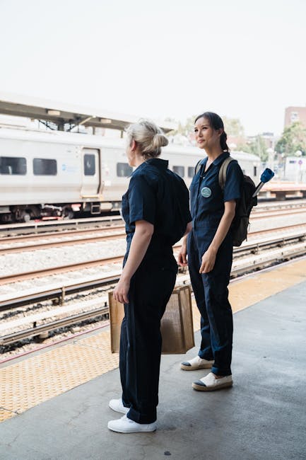 A person wearing a dark blue and orange jacket with 'ROLLINGSTOCK' written on the back, carrying a black backpack, is walking along a platform at a train station. The platform has a green railing on t