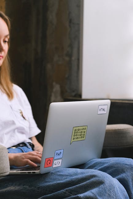 This image shows a person using a laptop, which is decorated with colorful stickers such as 'HTML', 'PHP', and a red power button icon, situated on their lap. The individual, with long brown hair, is