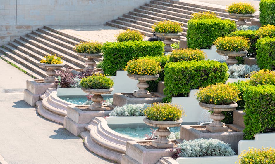 A landscaped garden features a series of wide, light-colored stone steps leading up through lush greenery, including neatly trimmed hedges and small bushes. To the right of the steps, there's a wooden